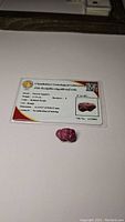 Natural untreated rough sapphire next to certification card from Chanthaburi Gemological Laboratory showing details including weight, color, cut, hardness, and no heat treatment
