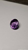 Faceted oval natural amethyst gemstone with deep purple color shown against plain background, highlighting size and clarity