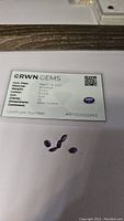 Photo showing six oval calibrated Bolivian amethyst gemstones placed on a white background alongside their authenticity certificate from GRWN GEMS, dated March 18, 2023, indicating Vs clarity, 6x4 mm dimensions, and purple color.