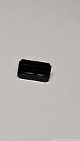 Close-up photo of oval-shaped dark olive green tourmaline gemstone showing octagonal faceting and polished surface under direct lighting on beige background.