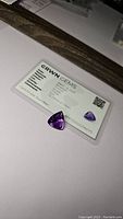 Photo showing the natural amethyst gemstone next to its certification card from GRWN GEMS with details: date March 18, 2023, 10.41ct weight, amethyst material, purple color, trillion cut, 15.73mm x 15mm dimensions