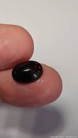 Close-up image of one jet black oval cabochon held between thumb and forefinger showing red play-of-color and polished smooth surface.