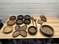 Large group of assorted vintage wooden bowls and serving utensils displayed on a wooden table against a white brick wall. Various sizes and shapes visible including round and divided bowls, large rustic bowl with bark edge, two long wooden spoons for salad serving, small spade-shaped piece, and smaller segmented dish.