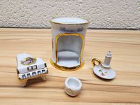 Four miniature porcelain items on wooden surface against textured wall: a grand piano, candle holder, chamber pot, and a holder with Bonne Nuit inscription.