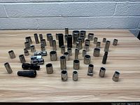 Photo showing assorted sockets arranged on wooden table in front of brick wall, various sizes and finishes.