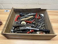 Top view showing assorted hand tools including pliers, hammer, clamps, utility knives in a wooden box