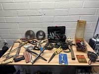 Wide view of assorted tools spread on a table against a brick wall, including saw blades, clamps, wires, pliers, and a toolbox.