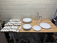 Photo of the complete lot showing stacks of white plastic plates in multiple sizes, rows of flatware wrapped in white napkins with grey rings, and clear plastic glasses.