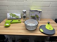 Photo showing the full lot of five kitchen items placed on a wooden table against a white brick wall. Includes salad spinner components, mandoline slicer and accessories.