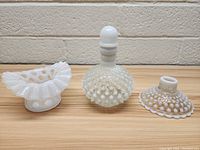 Photo showing three vintage white milk glass items including a hobnail perfume bottle with a rounded stopper and two hobnail lampshade parts placed on a wooden table against a brick wall background.