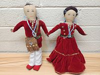 Pair of vintage fabric dolls wearing traditional styled clothing with bead and button decorations, standing against a wall.