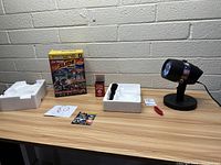 Star Shower LED Slide Show projector and accessories on a table against a white brick wall. Shows the projector, instruction manual, remote control, 12 slides, and packaging materials.