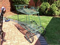 Side view of metal beverage cart showing large front wheels and mesh shelves with bottle holders on bottom.