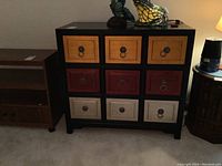 Front view of wood chest showing nine drawers with orange, burgundy, and cream fronts, black sides and top, metal ring pull handles.