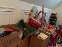 Multiple boxes filled with assorted Christmas items including wrapping paper rolls, gift bags, and garlands set against a white paneled wall.