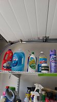 Wide view of upper shelf containing bottles of Resolve carpet shampoo, Ultra dish soap, Soft Scrub cleaner, Windex, Lysol spray, Clorox bleach, and other cleaning products.