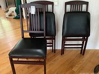 Four wood frame folding chairs with black vinyl seats and slatted backs visible, placed standing and leaning against wall on wood floor