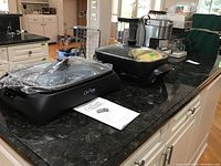 Chris Freytag Health Grill with clear glass lid, wrapped in plastic, on a kitchen counter with its instruction manual