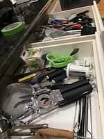 Two drawers filled with various kitchen utensils including strainers, whisks, spatulas, measuring cups, skewers, green plastic apple corer, mixer attachment, and knives visible.