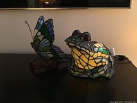 Both stained glass lamps together showing the glowing frog lamp and butterfly lamp on a dark surface.