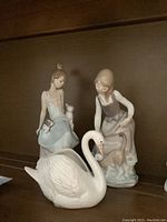 Three Lladró figurines on shelf: swan, seated girl with kittens, standing girl with cat