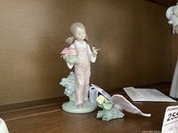 Front and side view of porcelain figurine showing girl with pink overalls holding bird and flowers standing next to a watering can on foliage base.