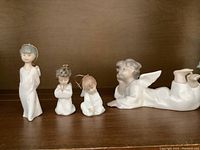 Four Lladro angel figurines on wooden shelf: three small with hanging loops, one larger lying down figure