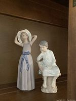 Two Lladro porcelain figurines on a wooden shelf, one girl standing with hat and dress, the other boy seated.