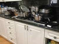 Full view of kitchen counter displaying multiple pots, pans, saucepans, and a rectangular stainless steel pan arranged around a stovetop.