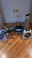 Overview of 5 electronic items on wooden floor: alarm clock radio, boom box, tower fan, space heater fan, boxed karaoke player with microphone.