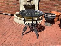 Full view of black wrought iron cafe table outdoors by brick fountain and planters.