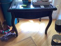 Full view of black rattan side table showing woven rattan legs and frame with glass top and some surrounding items not included