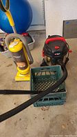 Photo showing side view of yellow Eureka upright vacuum cleaner and black Porter-Cable wet dry vacuum with hose on green crate.