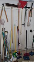 Wide shot showing assortment of long-handled yard and cleaning tools including hedge trimmers, rack rake, mop heads, brooms, and shovel standing against wall in garage.