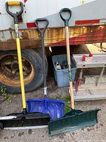 Three snow shovels leaning against a trailer and some equipment. Varied handles and blade styles visible.