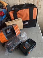 Photo showing the Ridgid branded carrying bag, battery charger, and two batteries wrapped in plastic on a table.