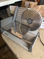 Photo of Cuisinart food slicer showing large circular metal blade, the metal food carriage, adjustable thickness control knob, and power cord on beige surface.