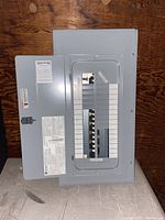 Gray electrical panel lying flat showing front with glass window and multiple breakers inside, labels and instruction stickers visible.