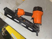 Photo showing an orange and black pneumatic framing nailer on a concrete floor, highlighting the air connector, magazine, and trigger handle.