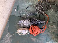 Two hanging work lights placed on a green surface, one with a black cord and one with an orange cord, both with metal cages around the bulbs.