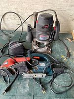 Three power tools on a workbench: Porter-Cable plunge router on top, Black & Decker jig saw and palm sander below, all with cords attached.