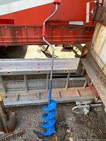 Full view of the manual post hole digger showing the red handle grip and the curved crank handle attached to a metal shaft above the blue auger blade.