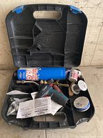 Open black plastic case with blue butane torch, black QLO gas lighter, solder wire rolls, metal nozzle, and instruction papers laid inside.