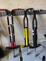 Photo showing three pogo sticks leaning against a wall with metal rack behind them. One red flame design, one yellow grip, and one black and white grip pogo sticks are visible.
