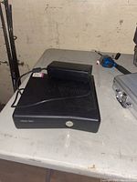 Black Xbox 360 gaming console on a table with a power supply brick resting on top. Console has visible ventilation slits and front power button.