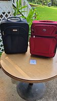 Photo showing two pieces of luggage on a round wooden table outdoors, one black and one red.