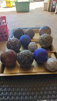 Photo showing eleven assorted decorative balls arranged on a gold-tone decorative tray. Balls vary in material with glass tile mosaics, embossed silver, blue textured, and grey rough finishes.