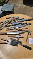 Photo showing collection of kitchen knives and utensils with wooden and synthetic handles laid out on a table outdoors