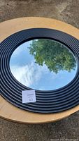 Full view of the round mirror placed on wooden table, showing black grooved plastic frame and clear reflective surface.