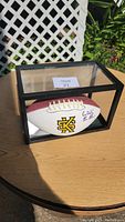 Signed Kansas State University football inside a clear display case on a wooden table outdoors.